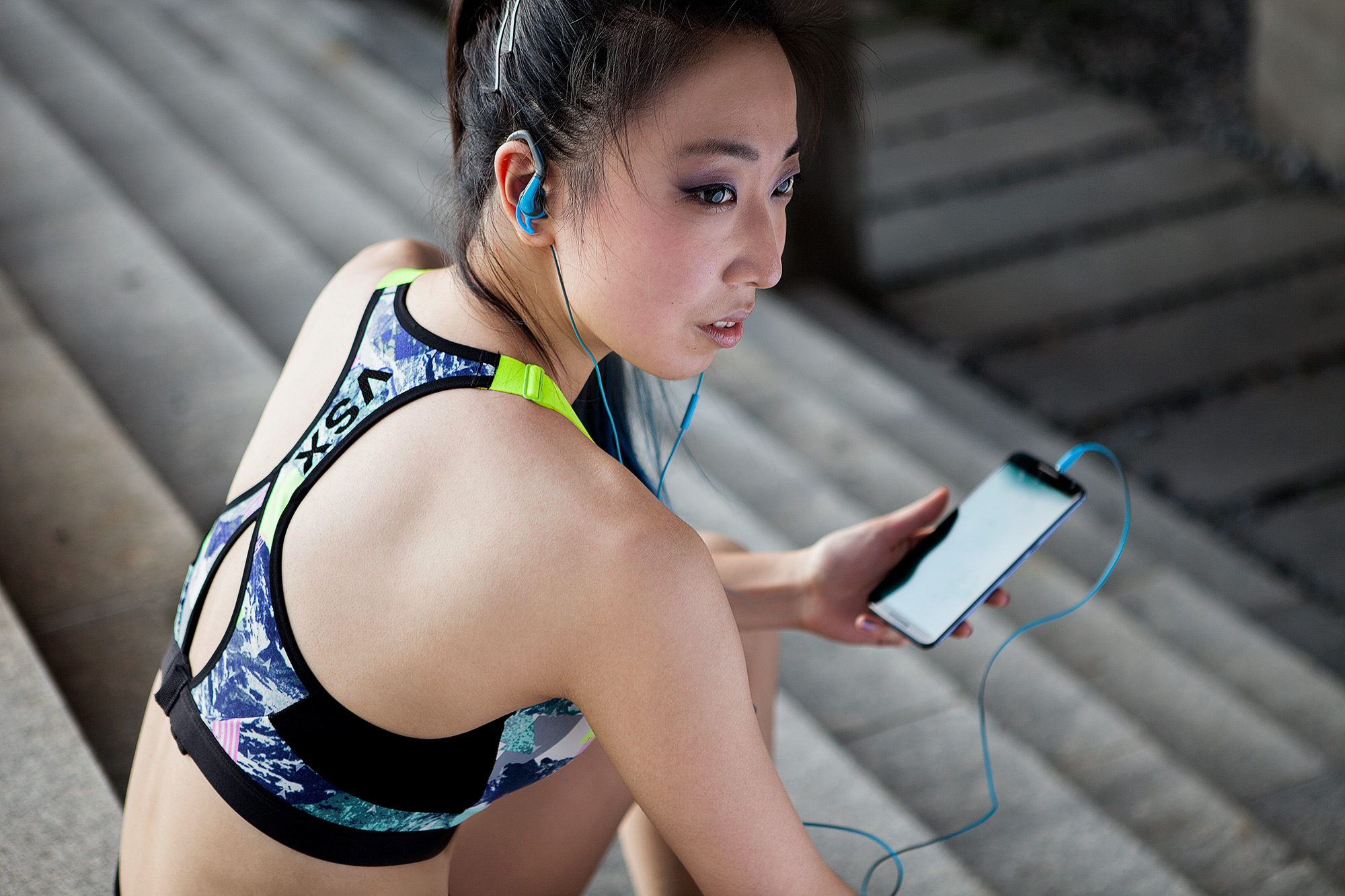Commercial fitness photography services A young woman wearing a sports top, listening to music on her phone while sitting on a staircase, artfully captured as part of a commercial fitness photography campaign.
