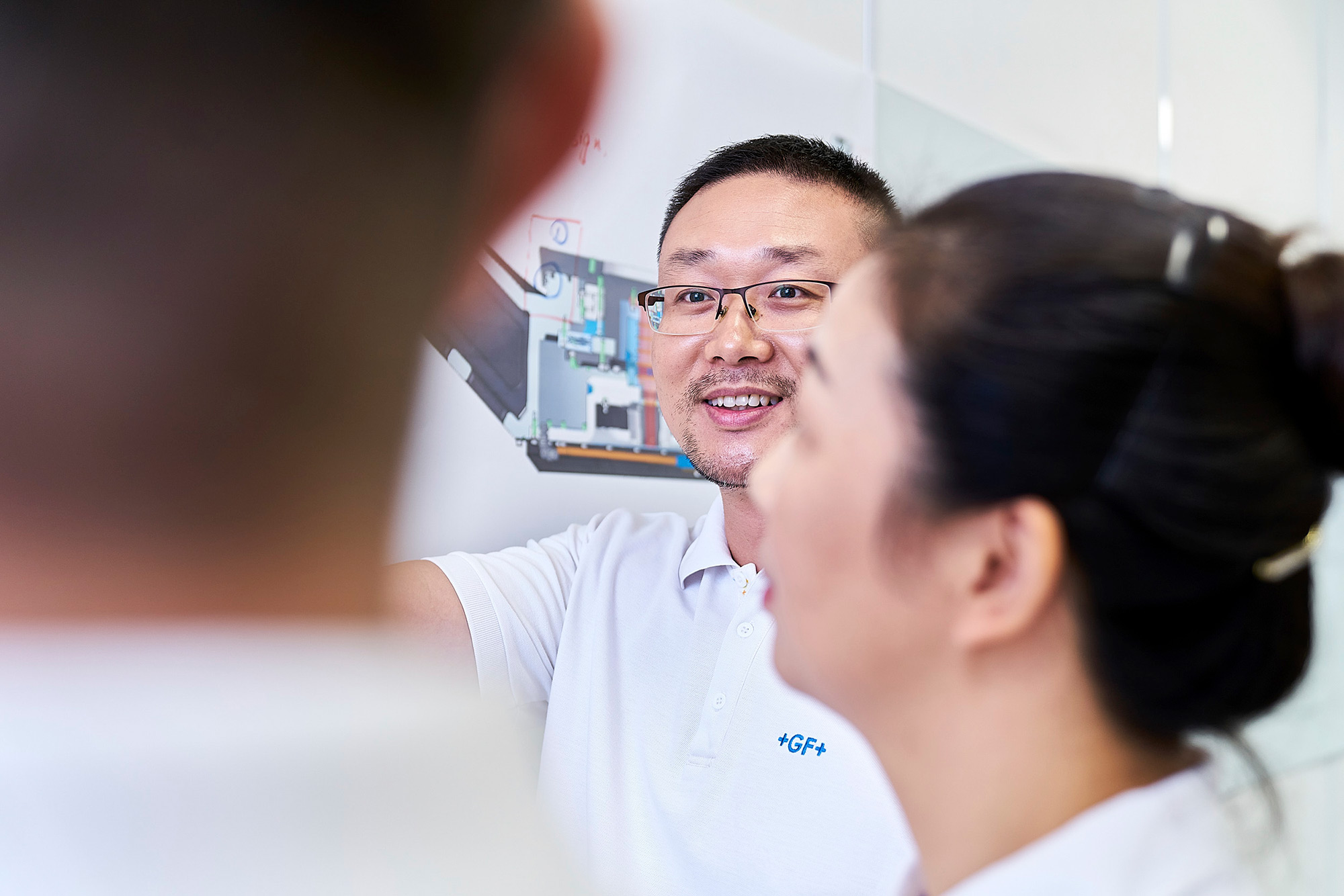 Corporate Photography Services. Three knowledgeable GF employees engage in a discussion about engineering solutions in front of a whiteboard with blueprints, captured in a corporate photography campaign.