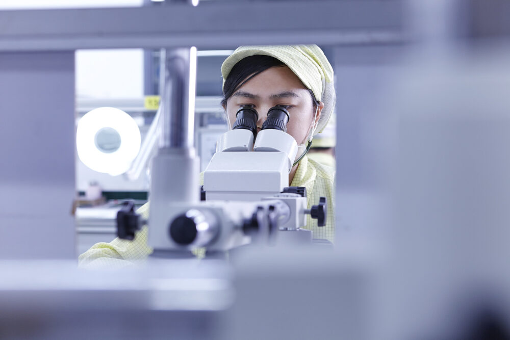 Female quality inspector examining through a microscope in a clean room, captured for Flextronics' science and technology photography campaign.